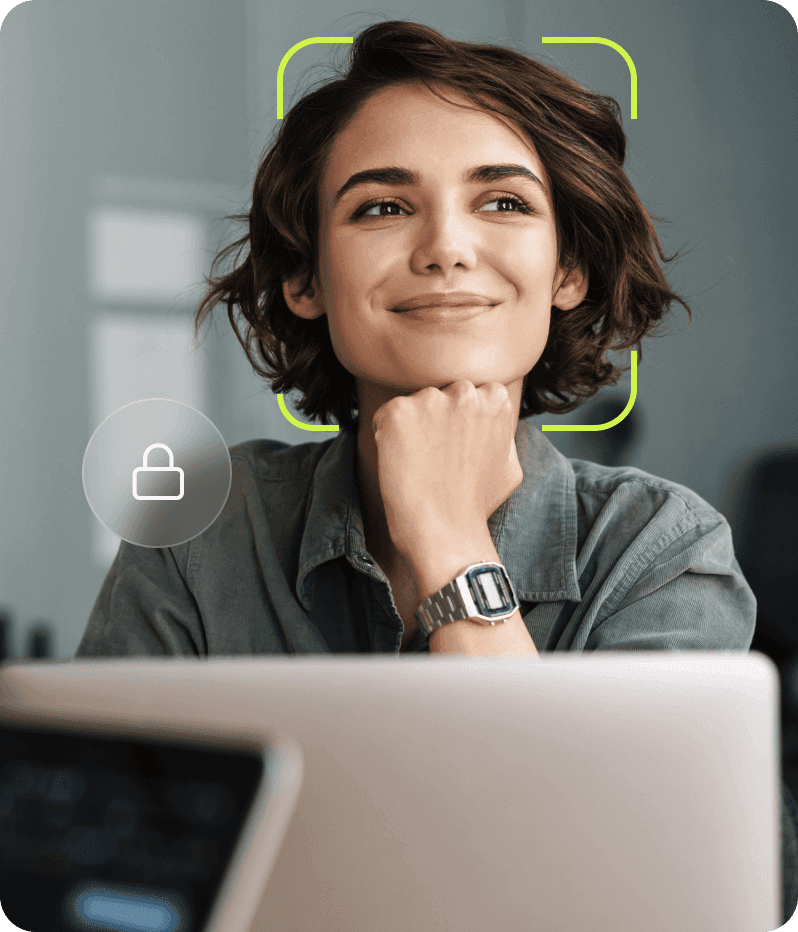Mulher jovem de cabelo curto sorrindo com o queixo apoiado na mão, com um retângulo de reconhecimento facial ao redor do rosto e um ícone de cadeado indicando segurança, em frente a um laptop em um ambiente de trabalho.